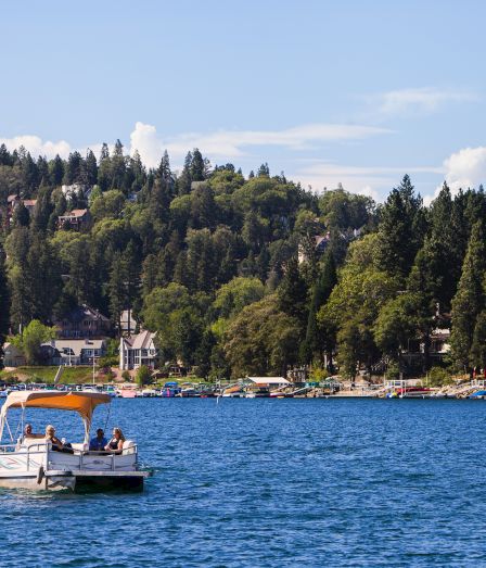 A small boat cruises across a lake toward a marina packed with yachts, with people on board and a forested hillside behind.