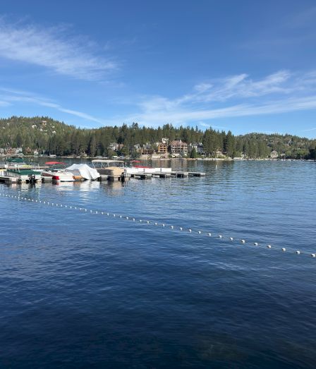 A calm lakeside marina with docked boats, floating platforms, hillside homes, and a blue sky with wispy clouds.
